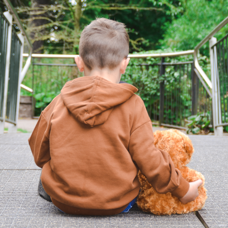 Little boy with brown jacket and back to camera holding brown teddy. Bereaved military child